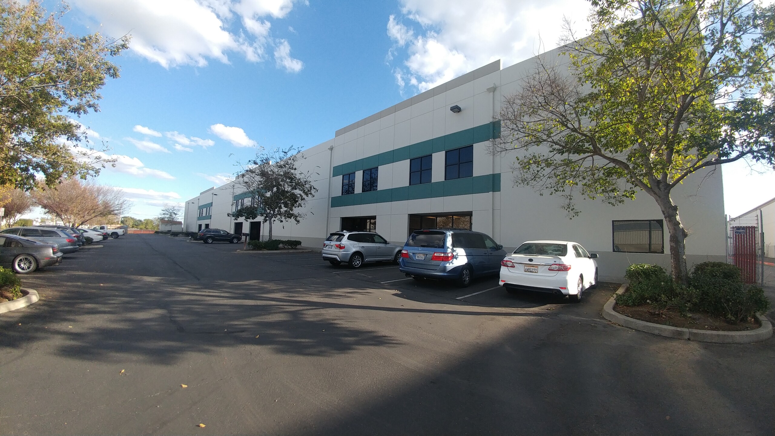 A wide-angle view of a modern industrial office building with a light beige facade and teal accent stripes. Several cars are parked in front of the building along a clean, paved parking lot. The scene is framed by a few trees, some with green leaves and others bare, under a partly cloudy blue sky.