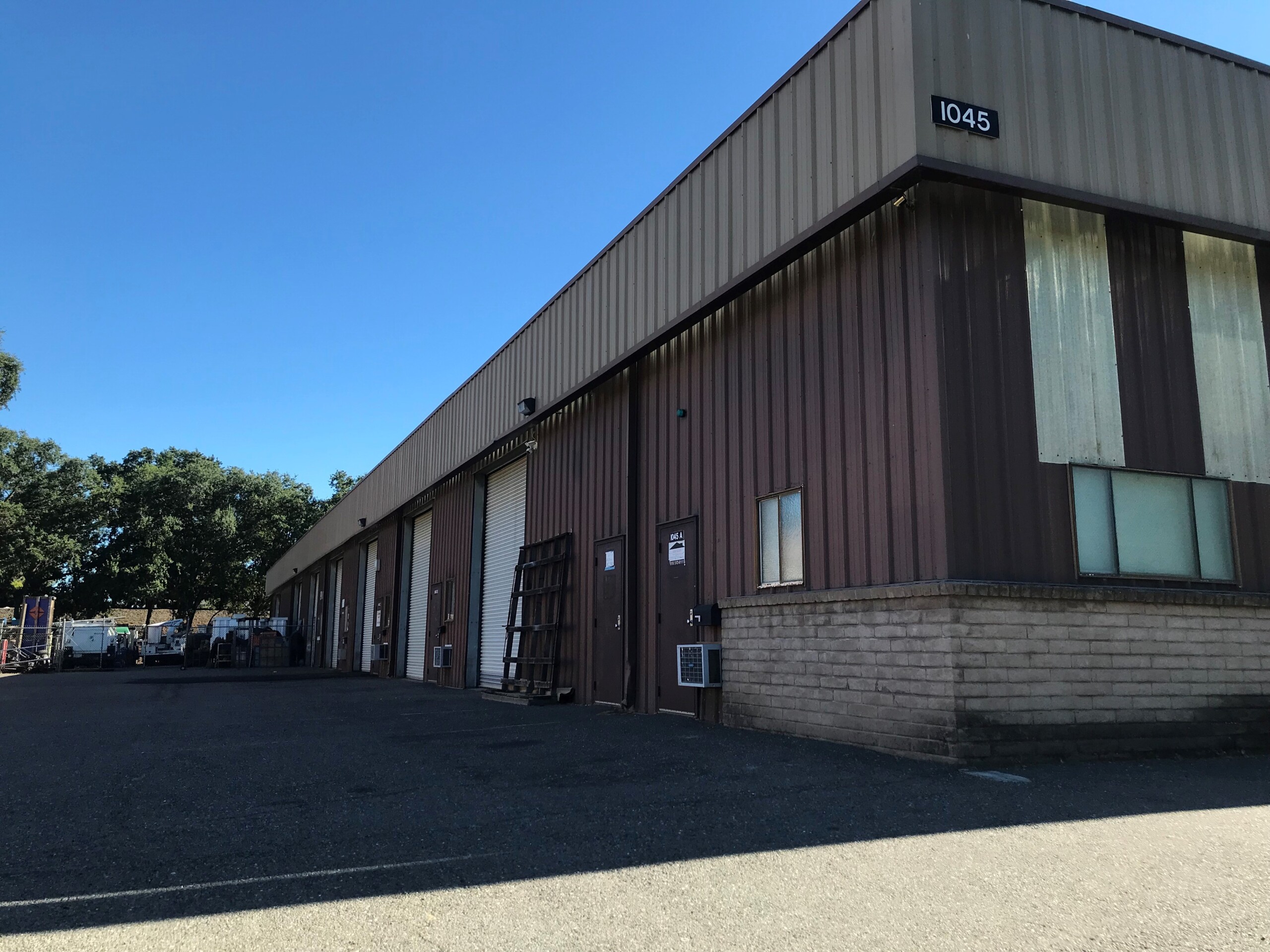 "Exterior view of a brown industrial warehouse building with multiple roll-up garage doors and entry doors. The building has metal siding with a beige trim and a sign displaying the number 1045 at the top corner. A paved lot runs along the front, and trees are visible in the background.