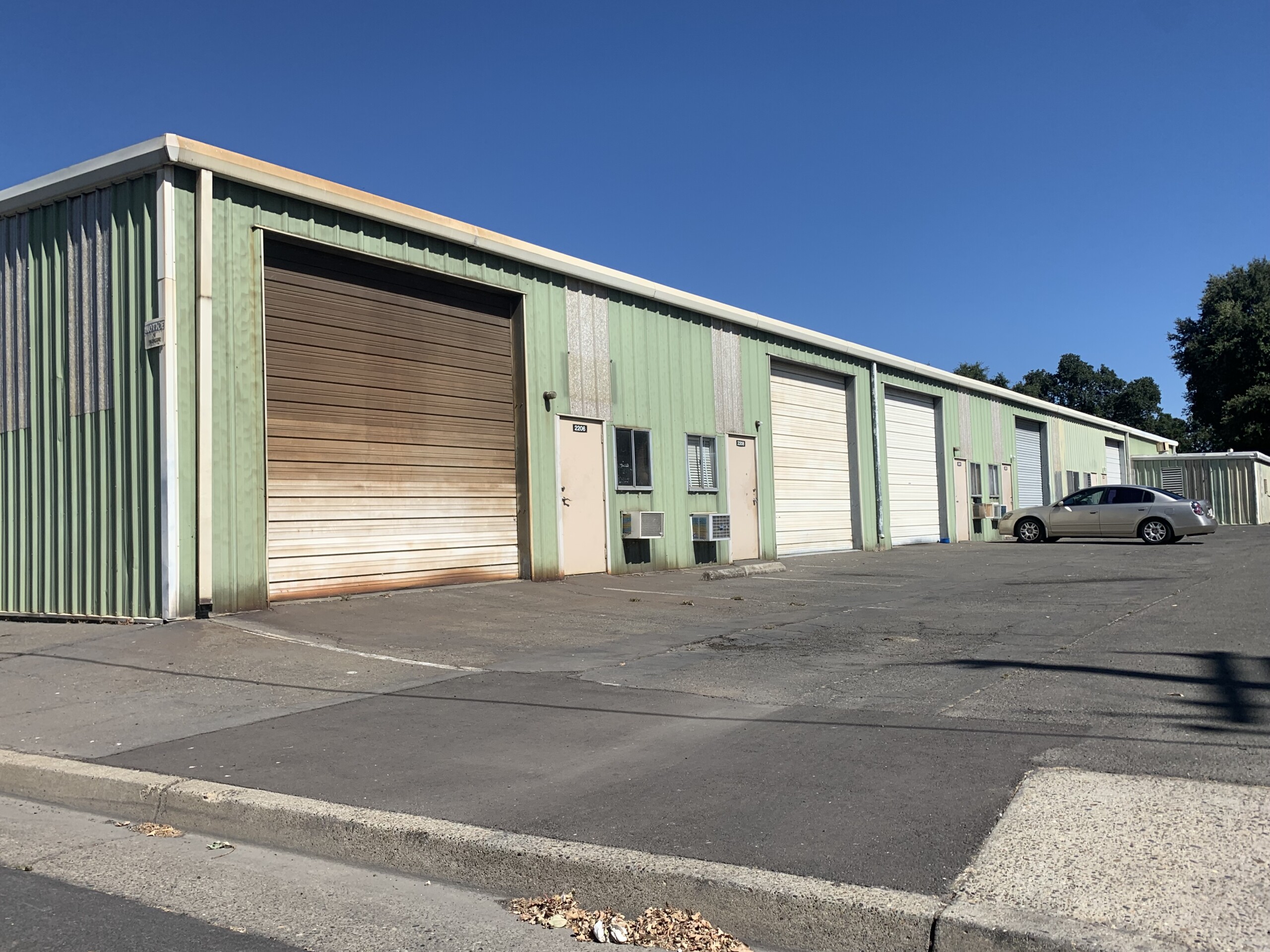 Single-story industrial warehouse building with green corrugated metal siding and multiple roll-up garage doors, some weathered with rust. A beige sedan is parked in front on the paved lot under a clear blue sky.