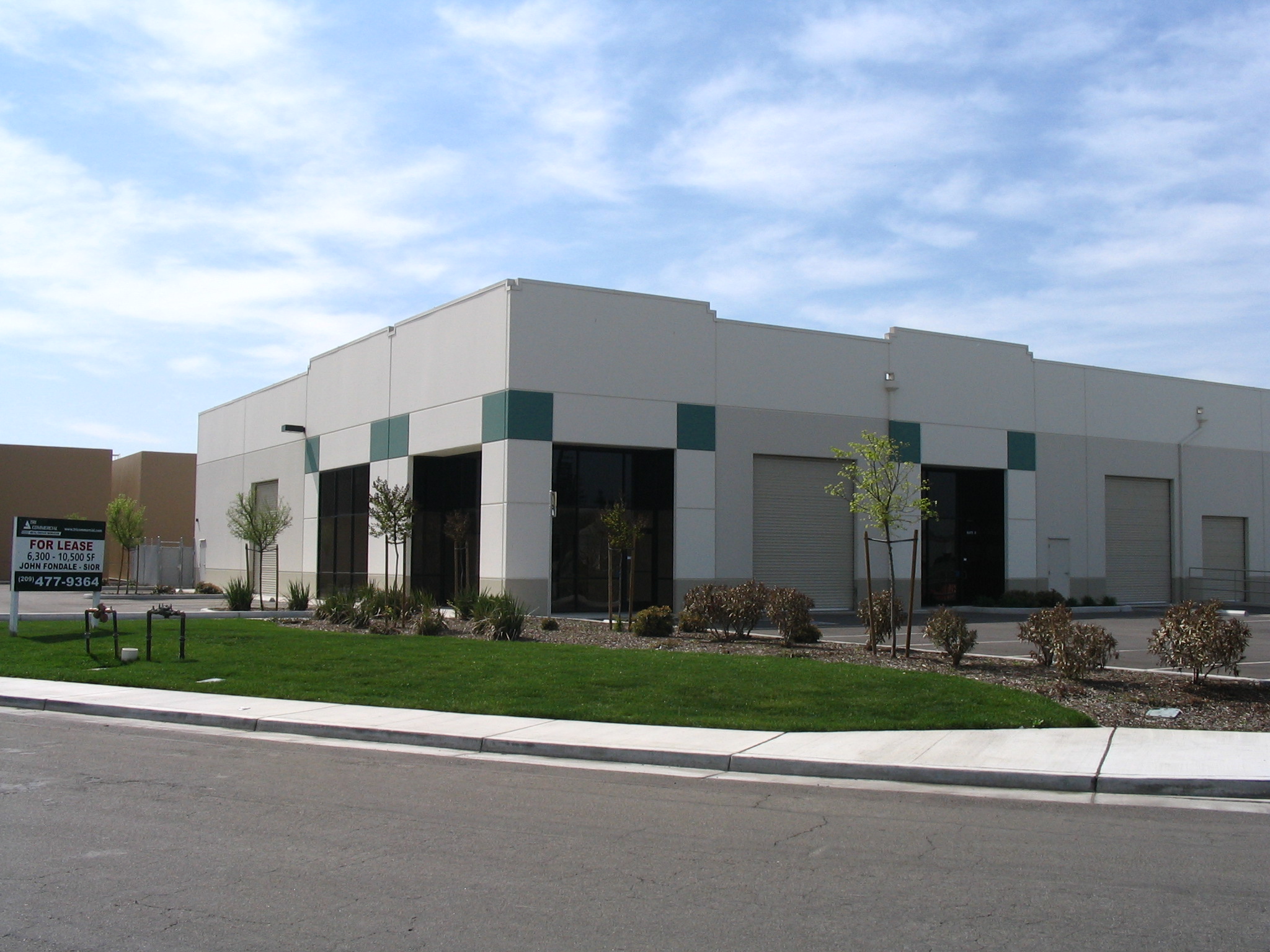 Modern single-story industrial warehouse building with multiple roll-up doors and office windows, landscaped with grass, young trees, and shrubs. A 'For Lease' sign is visible on the lawn near the street.