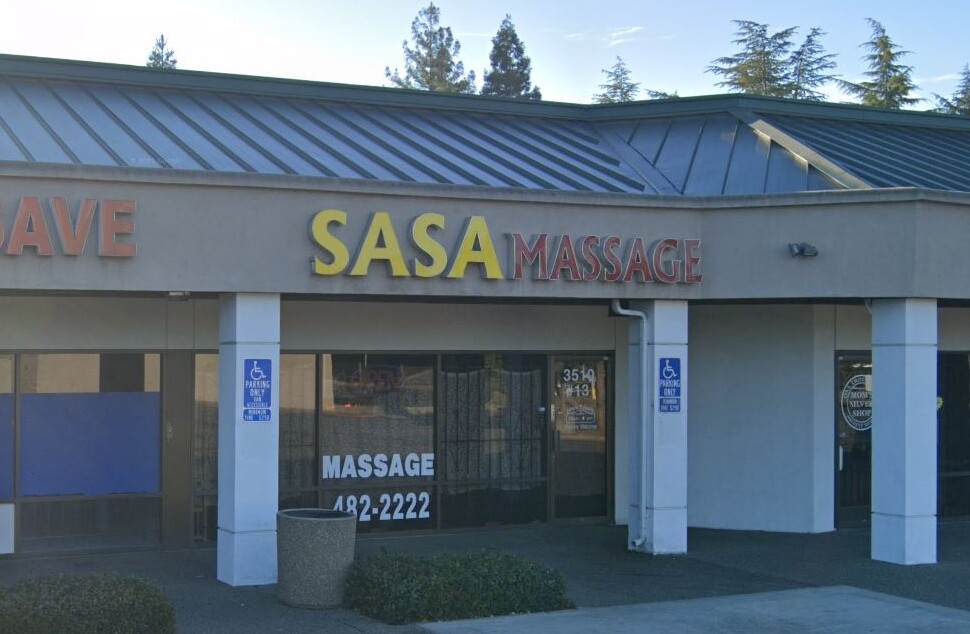 Street view of a small retail strip center. Visible businesses include a store with a sign reading 'PIK & SAVE,' a massage business with the sign 'SASA MASSAGE,' and another shop with a circular sign reading 'MOM'S SILVER SHOP.' The storefronts have large glass windows, and two handicap parking signs are posted near the entrances.