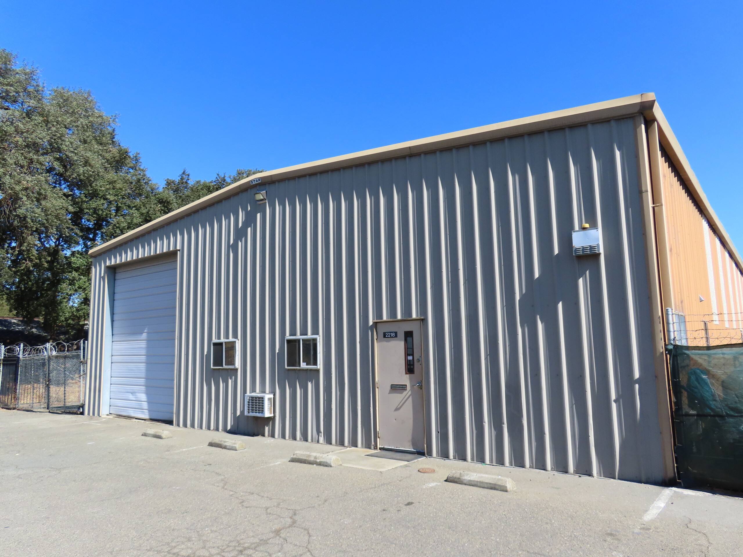 A light gray metal industrial warehouse with a roll-up door, two small windows, and a single entry door. The building sits on a paved lot with a few parking spaces and is surrounded by fencing. The sky is clear and blue with trees visible in the background.