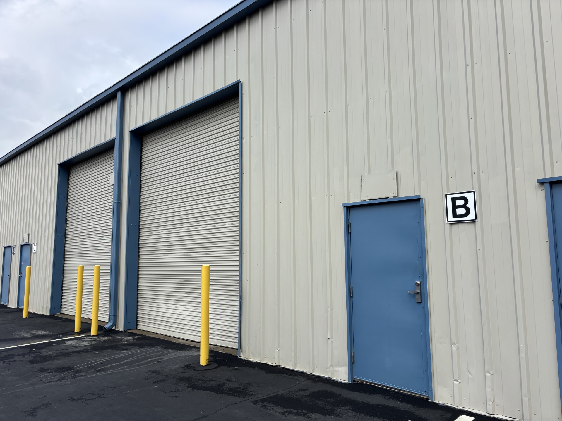 Exterior view of an industrial warehouse building with light beige metal siding and blue trim. The image shows two large roll-up doors, several yellow safety bollards, and a blue personnel door labeled ‘B.’ The asphalt driveway in front of the units is clean and well-maintained.