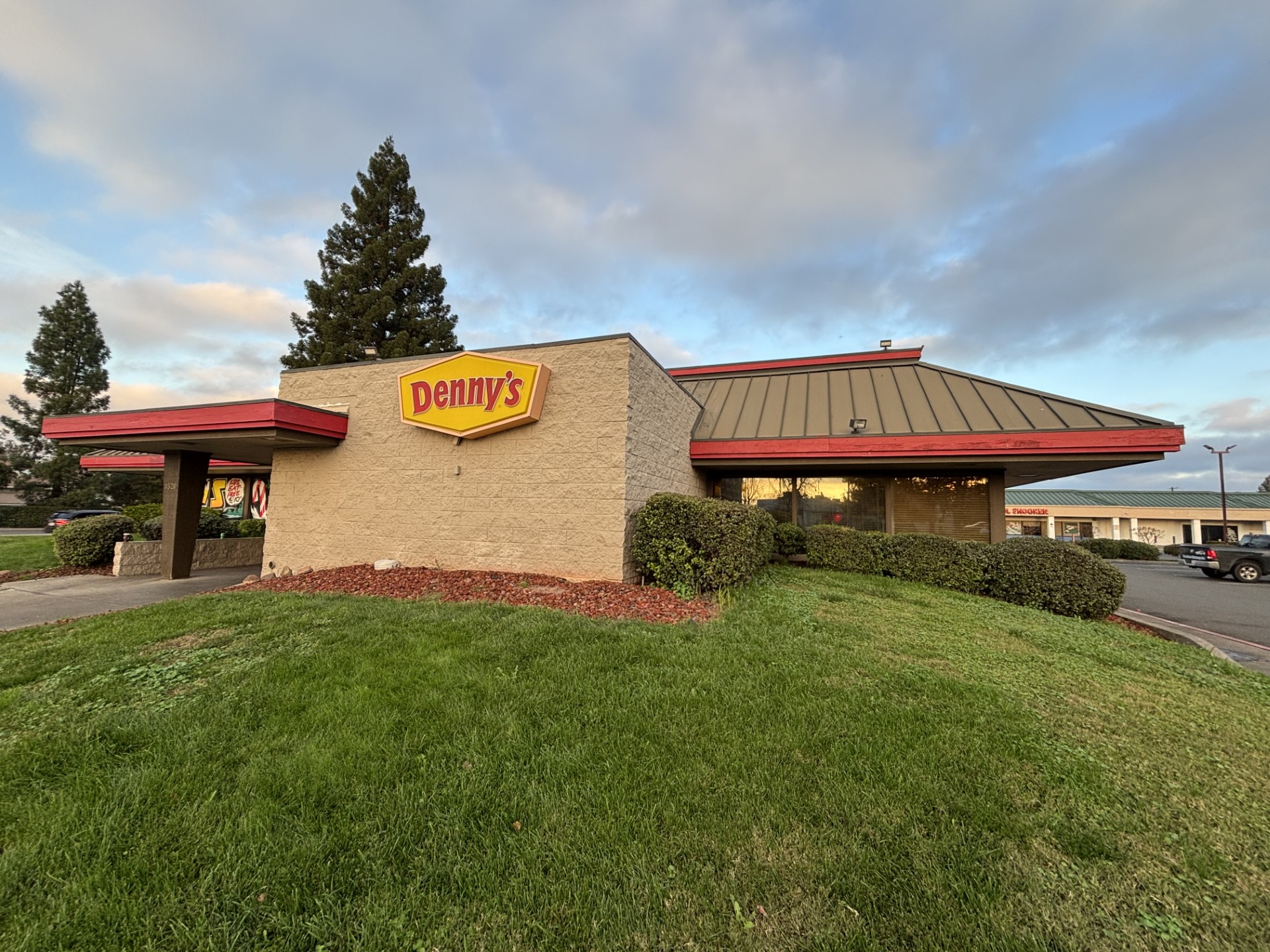 Exterior view of a single-story Denny’s restaurant featuring beige masonry walls, a red-trimmed roof, and the yellow Denny’s logo sign on the front facade, with landscaped shrubs, green grass, and a partly cloudy sky.